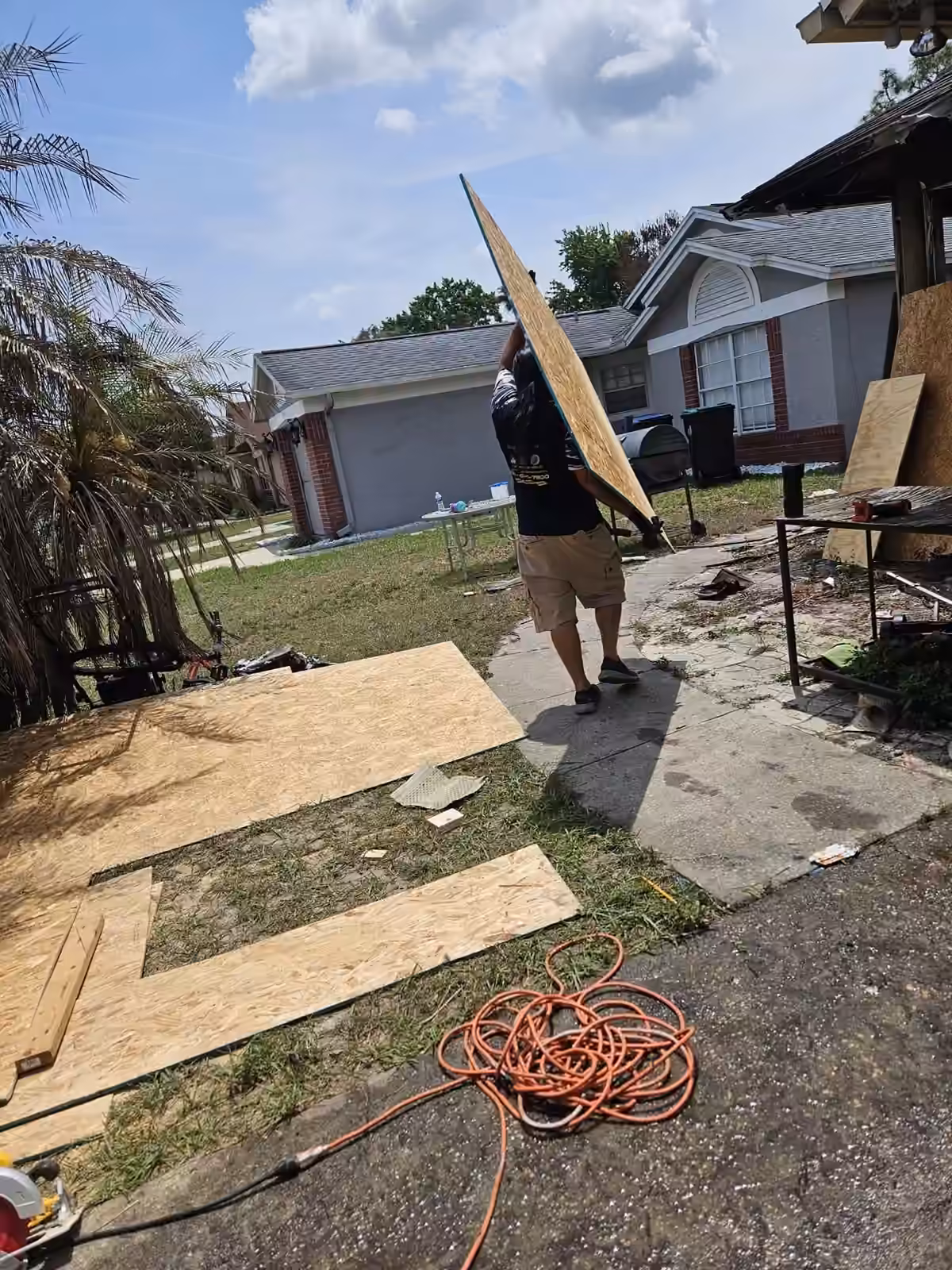 Travayè k ap pote yon panèl plywood deyò yon kay ki andomaje